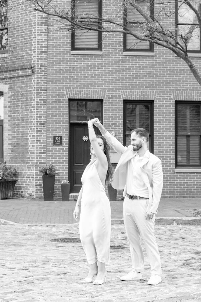 Baltimore wedding photographer captures a black-and-white photo of a couple twirling on the cobblestone streets of historic Fells Point during their engagement session.