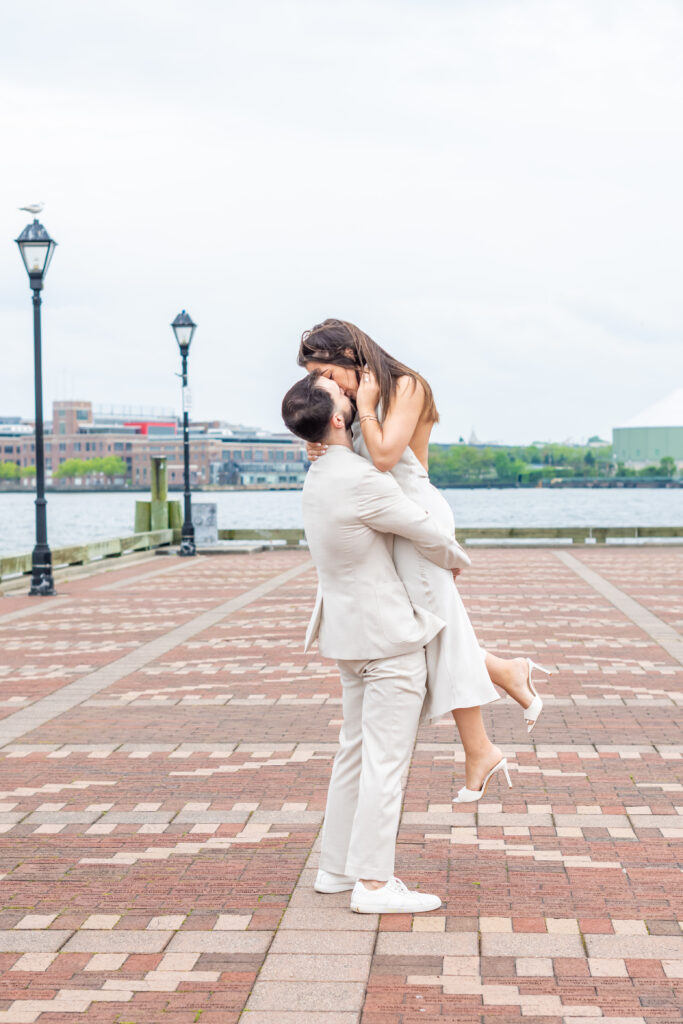 Baltimore wedding photographer captures a romantic moment during a Fells Point engagement session as the groom lifts his fiancée into a kiss along the brick waterfront promenade.