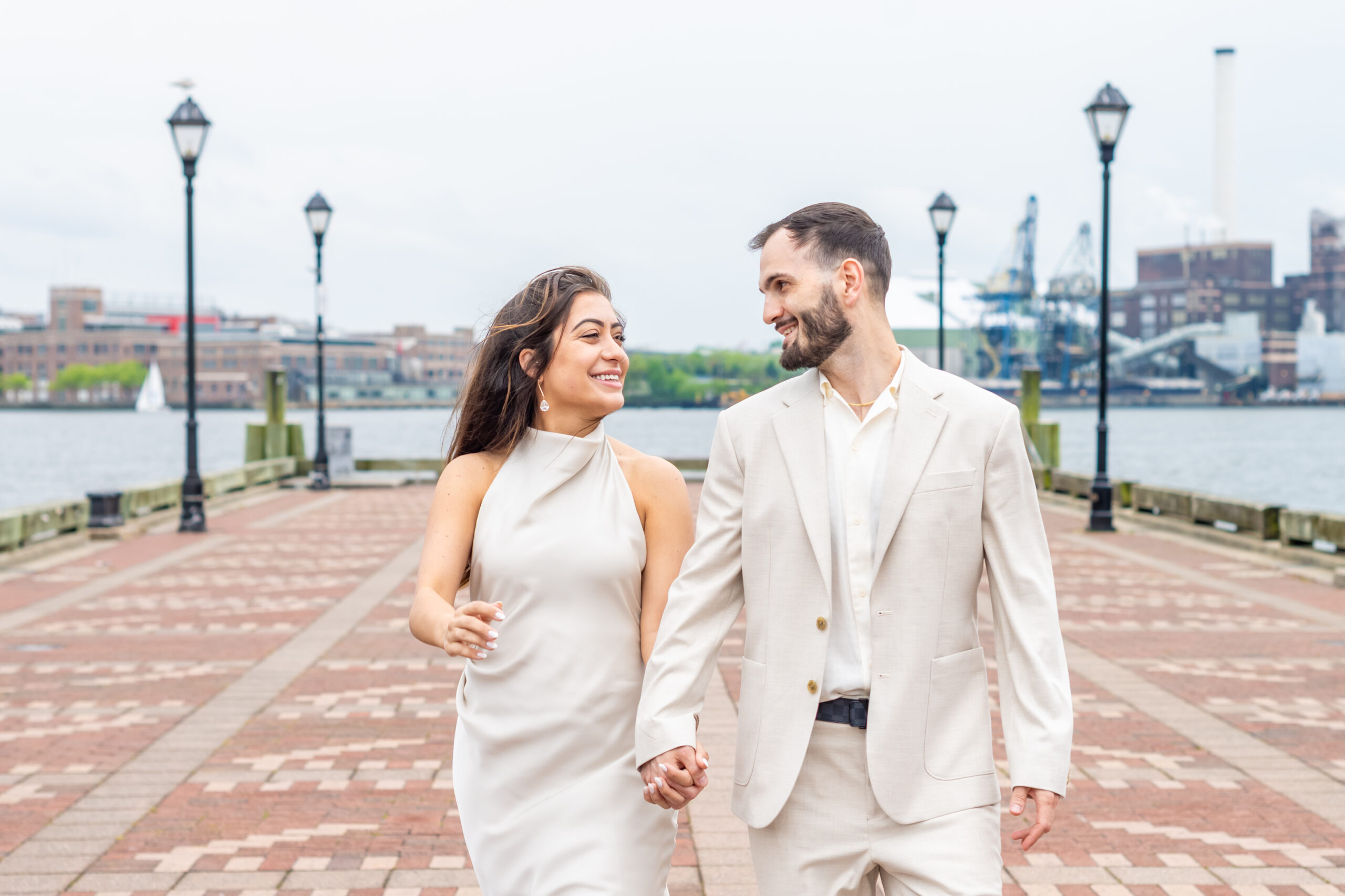 Baltimore wedding photographer captures a couple walking hand in hand along the Fells Point waterfront during their engagement session, smiling at each other against the scenic harbor and city skyline backdrop.