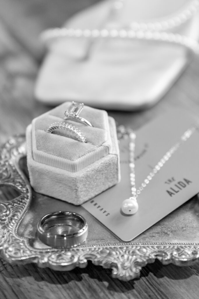 Elegant wedding rings displayed on a velvet ring box atop an ornate silver tray, paired with a pearl necklace and groom's band.