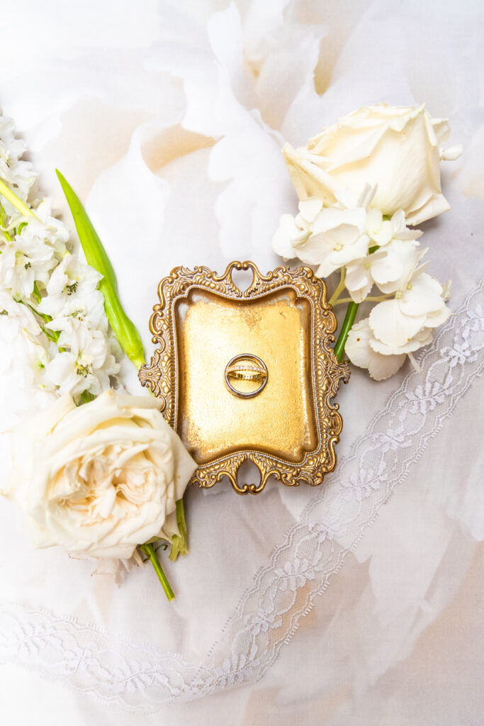 Gold ornate tray holding two wedding rings, styled with white roses and lace on a soft neutral fabric.