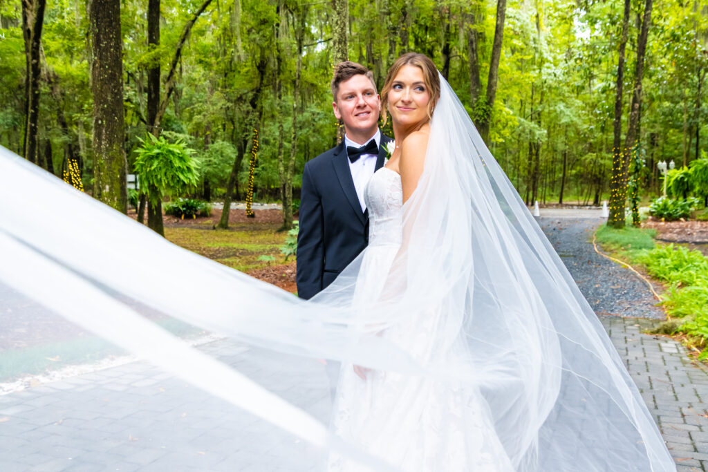 Bride’s veil flows in the foreground as she stands with her groom on a tree-lined path at The Mackey House.