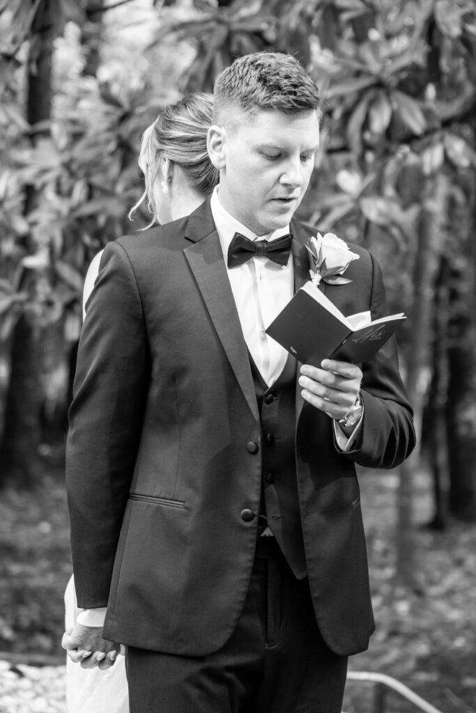 Groom reads from his vow book while holding hands with his bride behind him, captured in black and white.