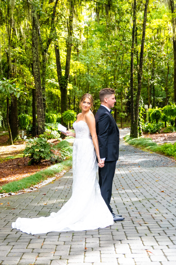 Bride and groom stand back-to-back holding hands while reading vows in a forested pathway.