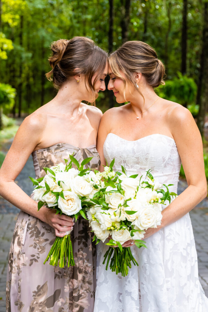 Bride and maid of honor stand forehead-to-forehead holding white bouquets on a tree-lined path at The Mackey House.