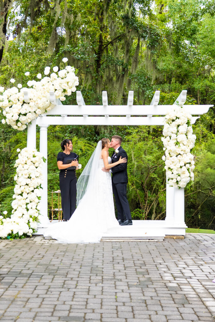 Bride and groom share their first kiss under a white floral arbor at The Mackey House.