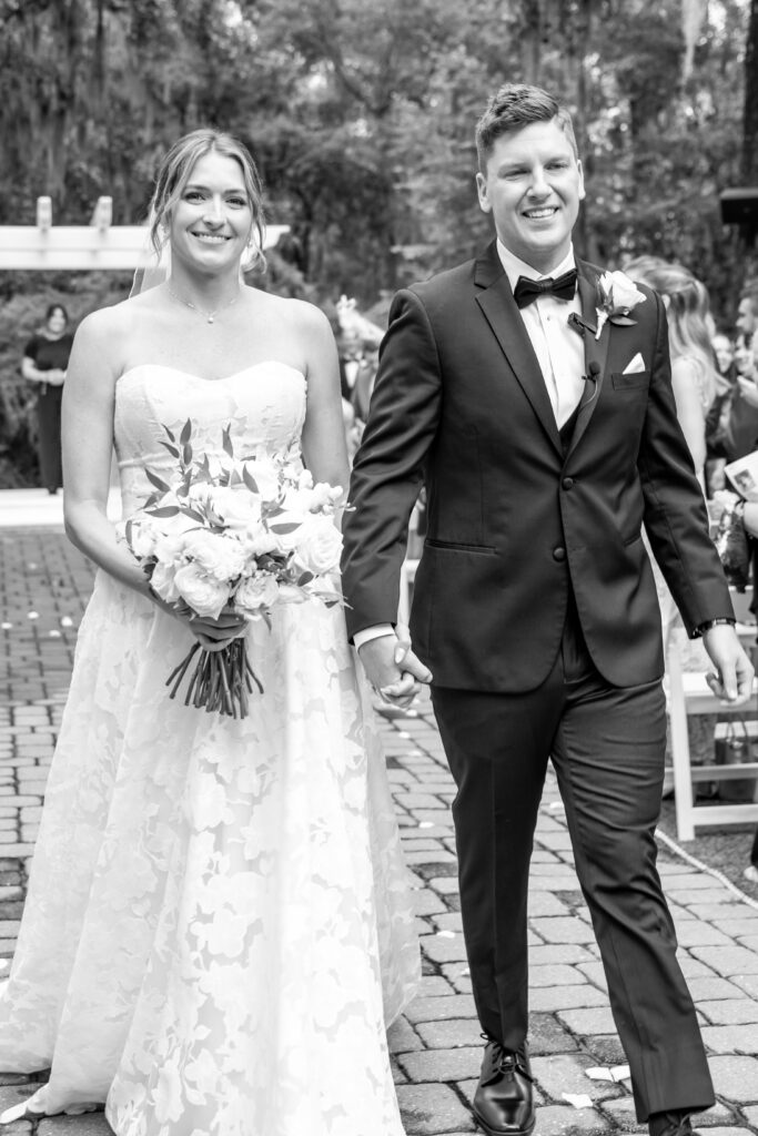 Black and white photo of the bride and groom smiling as they walk hand-in-hand up the aisle.