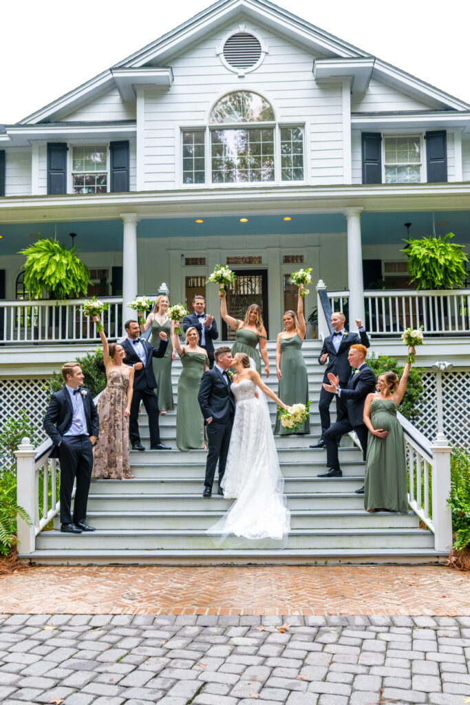 Bride and groom kiss on the staircase while their wedding party cheers behind them at The Mackey House.