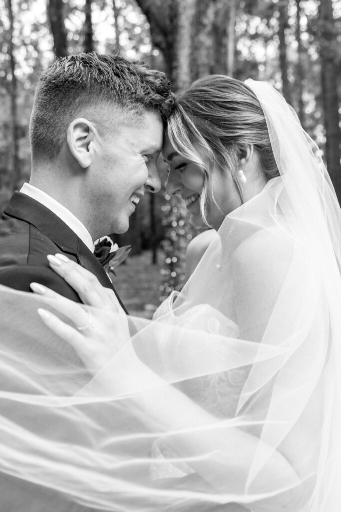 Close-up black-and-white photo of the bride and groom smiling forehead-to-forehead under the bride’s veil.