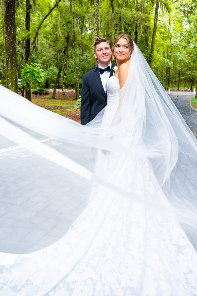 Bride’s veil flows in the foreground as she stands with her groom on a tree-lined path at The Mackey House.