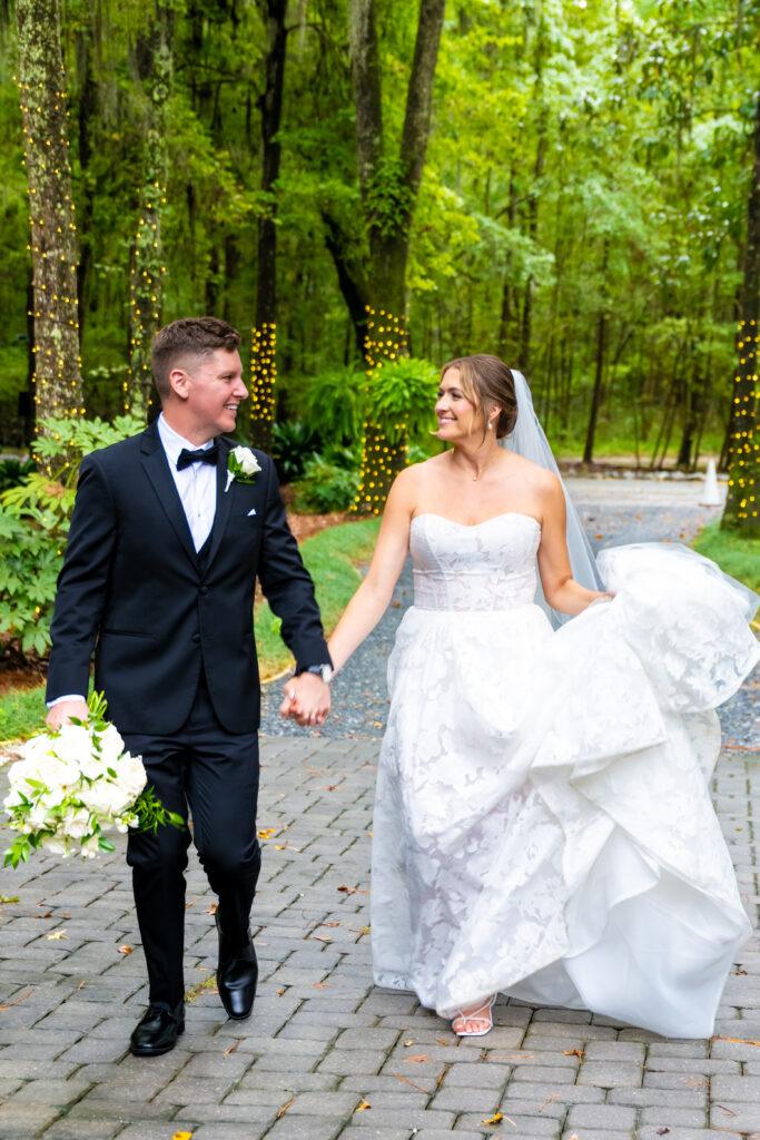 Bride and groom walk hand-in-hand down a wooded path, smiling at each other while holding the bouquet and dress.