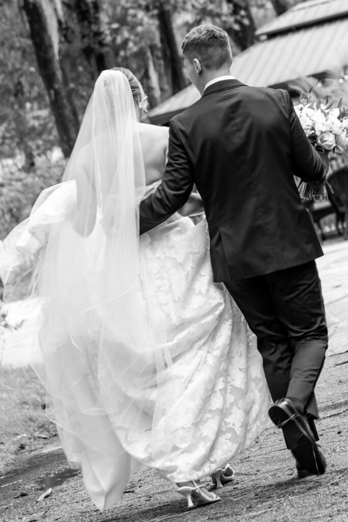 Bride and groom walking together after the ceremony, holding the wedding dress and bouquet, captured from behind.
