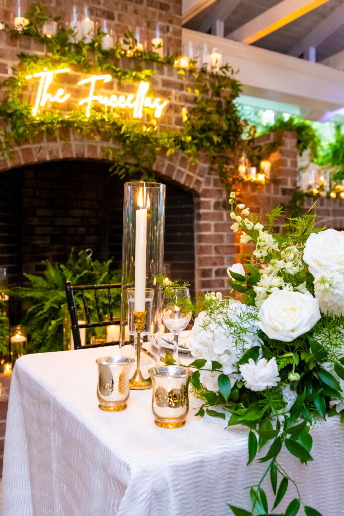 Elegant sweetheart table setup with white florals, candles, and a neon sign reading “The Fuccellas” above the brick fireplace.