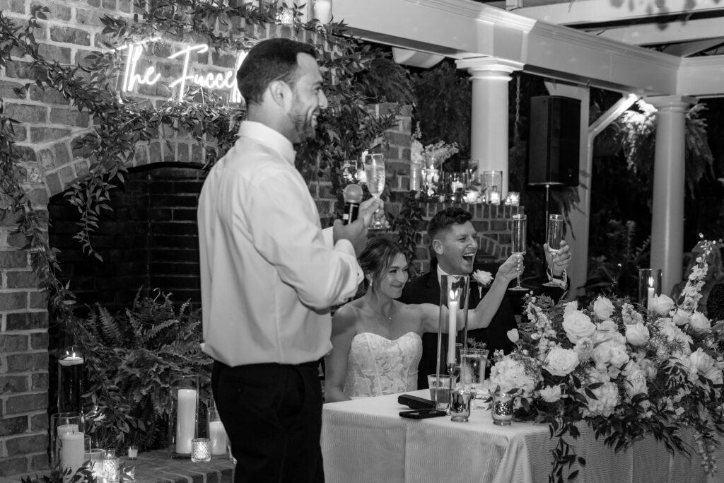 Best man giving a toast while the bride and groom raise their champagne glasses at the sweetheart table.