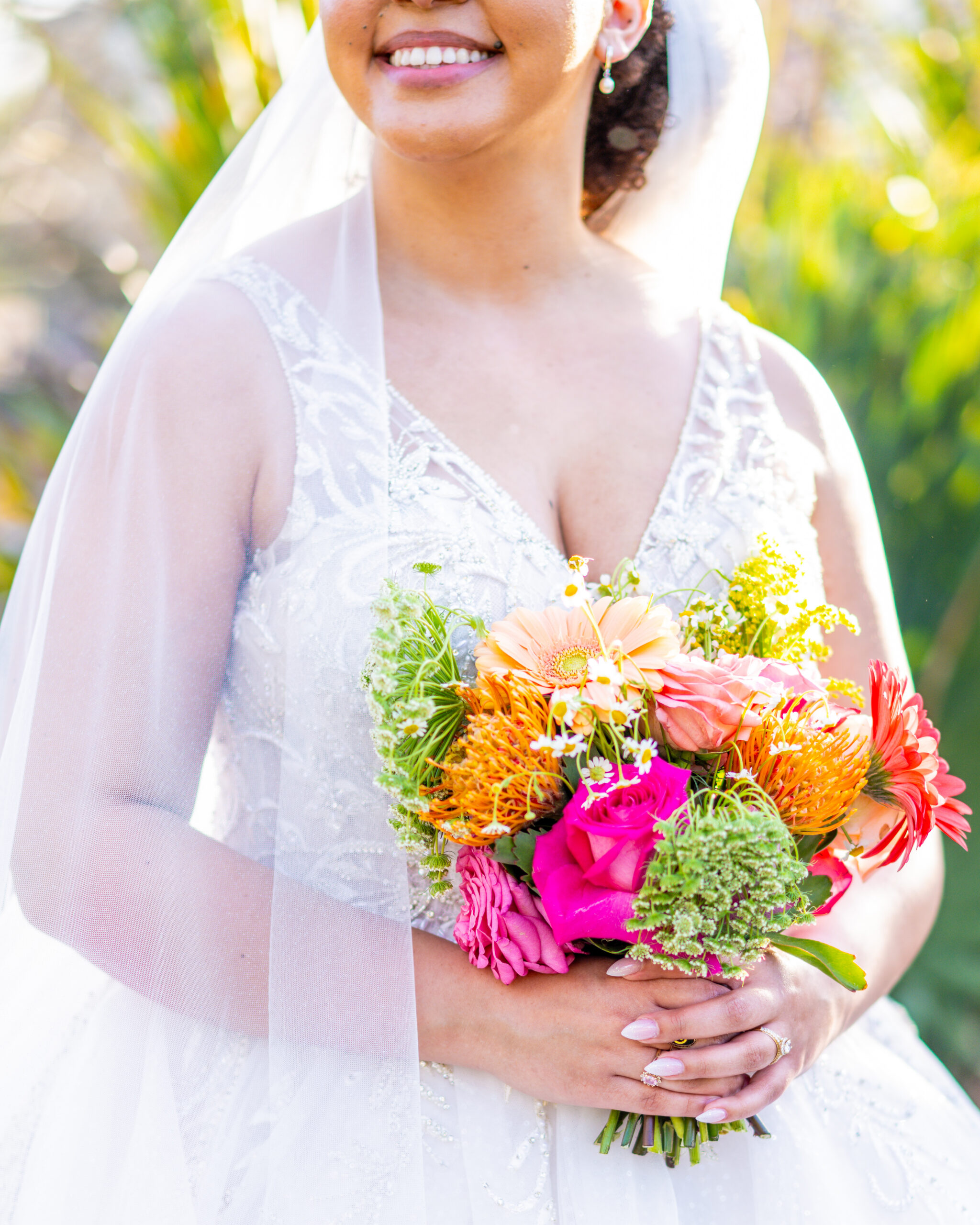 Smiling bride in a beaded lace gown and veil holding a vibrant bridal bouquet with orange gerbera daisies, hot pink roses, protea, and yellow wildflowers, photographed by Maryland wedding photographer Jennifer Mary Collective
