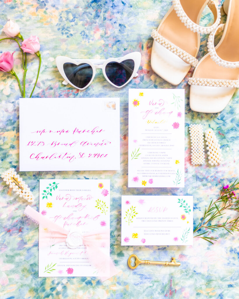 Colorful watercolor floral wedding invitation suite flat lay with hot pink calligraphy envelope addressing, pearl bridal shoes, heart shaped sunglasses, and pearl hair clips on a floral background, photographed by Maryland wedding photographer Jennifer Mary Collective