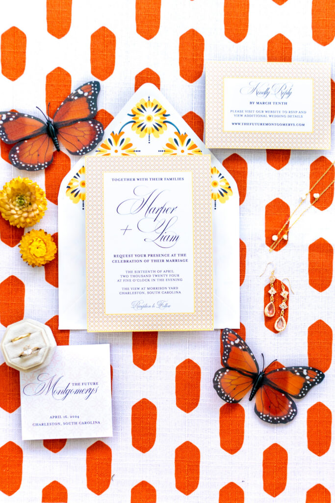 Bold wedding invitation suite flat lay on an orange patterned background featuring a geometric patterned invitation with yellow tile envelope liner, monarch butterfly accents, and gold jewelry, photographed by Maryland wedding photographer Jennifer Mary Collective