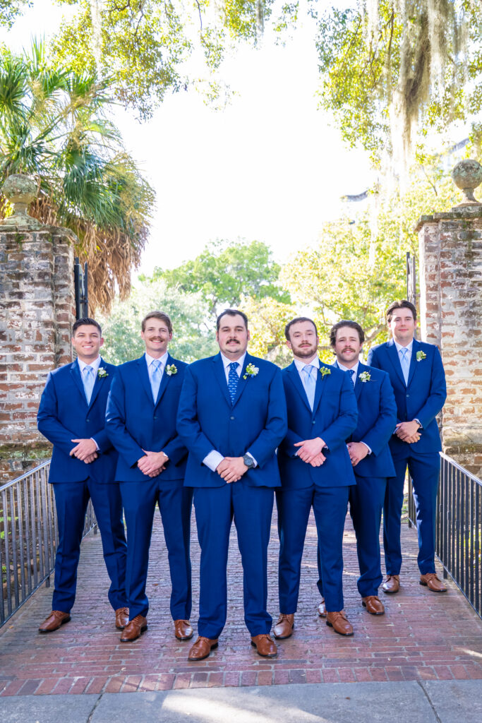 Six groomsmen in navy suits with white boutonnieres standing at a historic brick gate surrounded by trees, photographed by Baltimore wedding photographer Jennifer Mary Collective