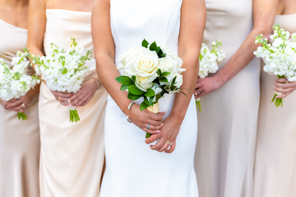 Close up of bride holding a white rose and greenery bouquet flanked by bridesmaids in champagne gowns holding white stock flower bouquets, photographed by Maryland wedding photographer Jennifer Mary Collective