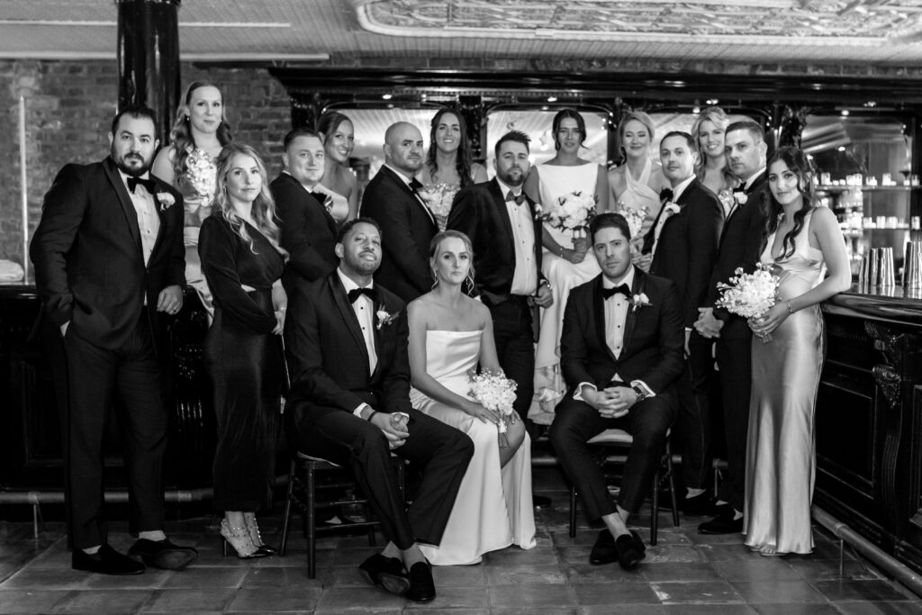 Black and white editorial group portrait of a large wedding party in black tie attire inside a beautifully ornate venue bar, photographed by Baltimore wedding photographer Jennifer Mary Collective