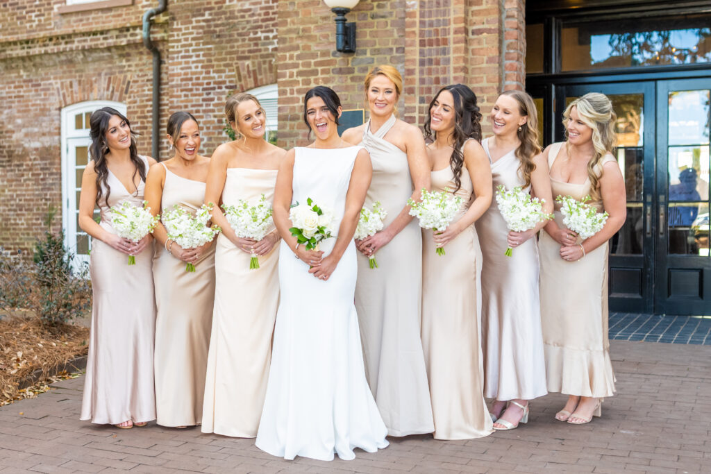Bride facing her bridesmaids in champagne satin gowns holding white floral bouquets outside a historic brick building in Annapolis, photographed by Baltimore wedding photographer Jennifer Mary Collective
