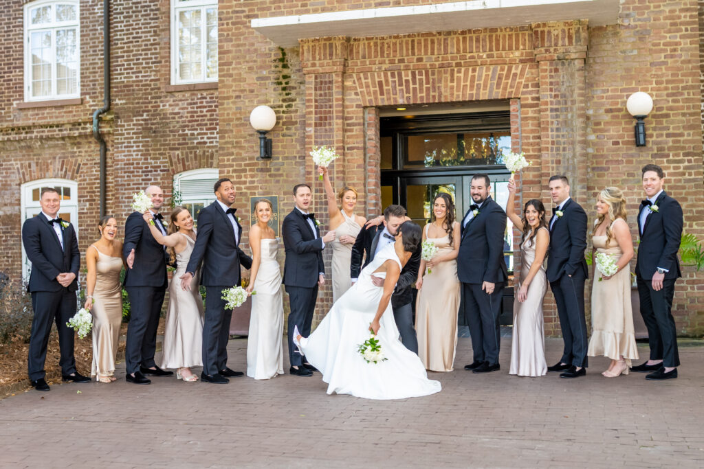 Groom dipping bride in a dramatic kiss while their full wedding party cheers and reacts joyfully outside a historic brick venue in Annapolis, photographed by Baltimore wedding photographer Jennifer Mary Collective