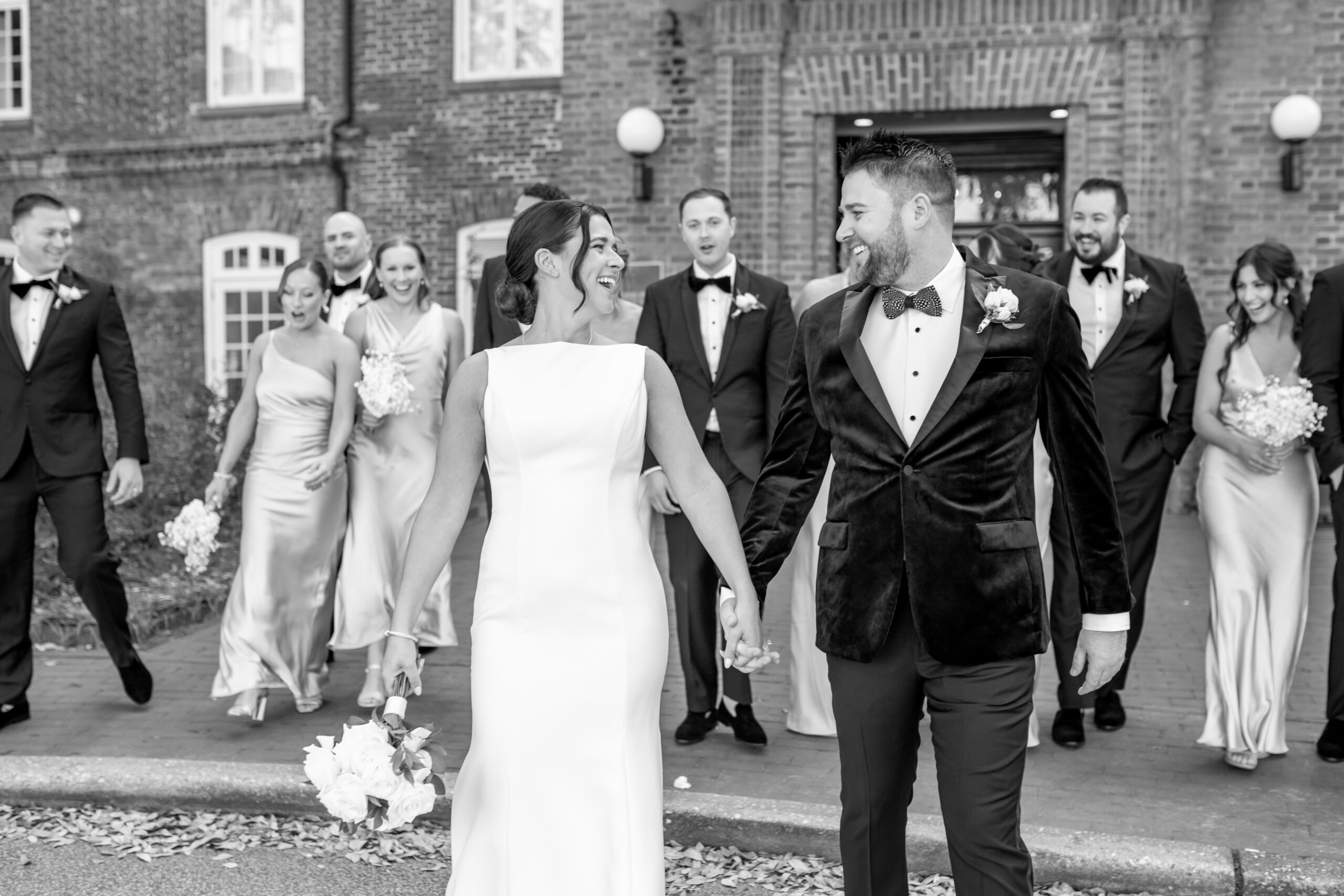 Black and white photo of bride and groom laughing while holding hands as their full wedding party walks behind them outside a historic brick building in Annapolis, photographed by Baltimore wedding photographer Jennifer Mary Collective