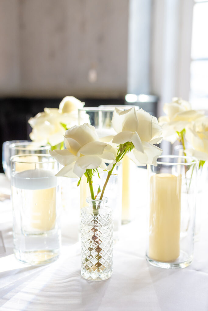 White roses in a crystal bud vase surrounded by pillar candles on a white linen reception table, photographed by Maryland wedding photographer Jennifer Mary Collective