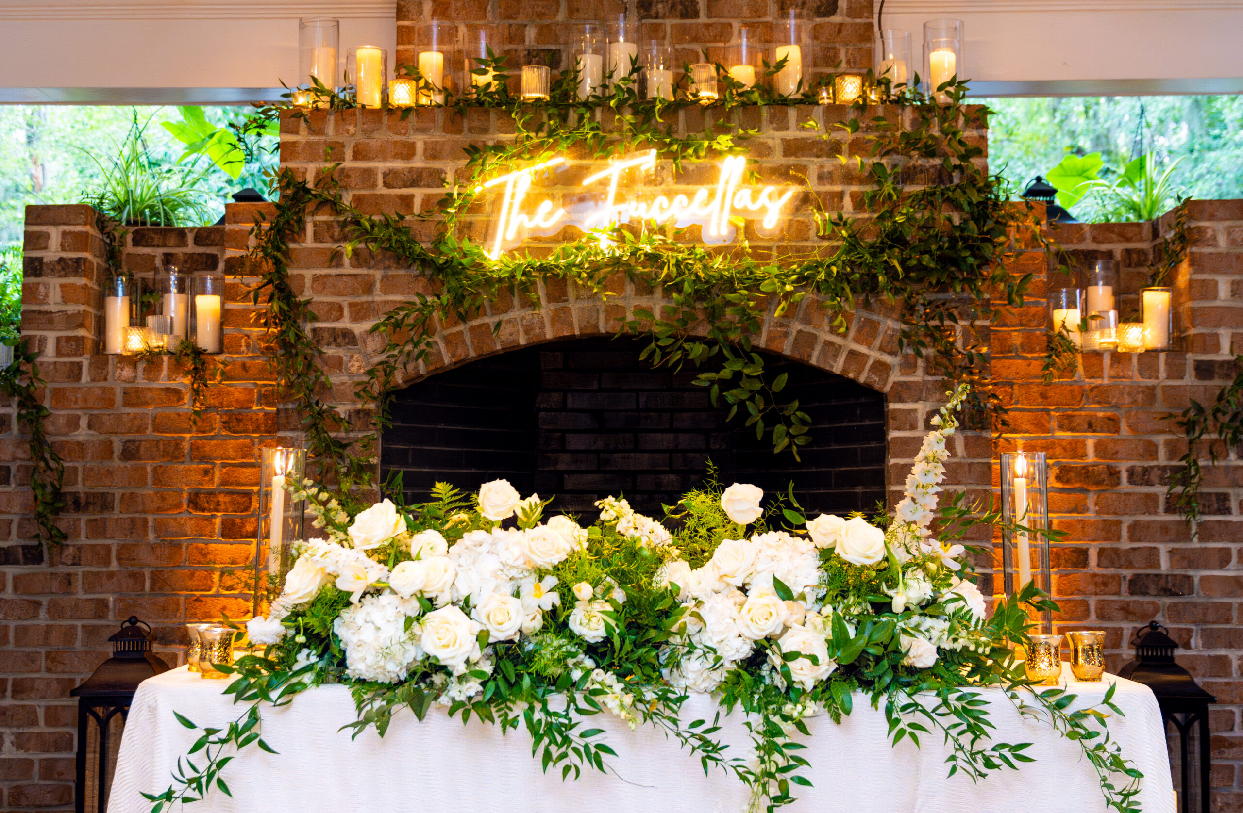 Wedding sweetheart table with a lush white rose and greenery floral installation in front of a brick fireplace decorated with garland, candles, and a glowing custom neon sign reading The Truccellas, photographed by Maryland wedding photographer Jennifer Mary Collective