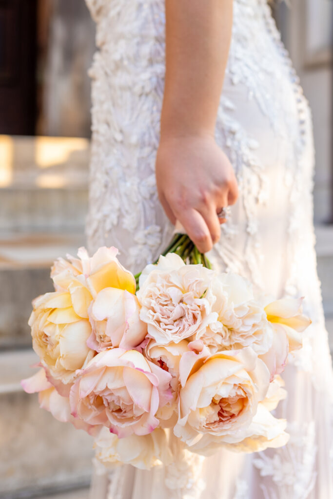 Bride in a heavily beaded lace gown holding a loose blush and cream garden rose bouquet at her side against warm stone steps in Baltimore, photographed by Baltimore wedding photographer Jennifer Mary Collective