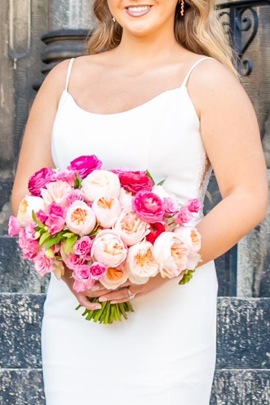 Bride in a simple white slip gown holding a vibrant bouquet of hot pink, blush, and peach garden roses in front of historic stone steps in Mount Vernon Baltimore, photographed by Baltimore wedding photographer Jennifer Mary Collective
