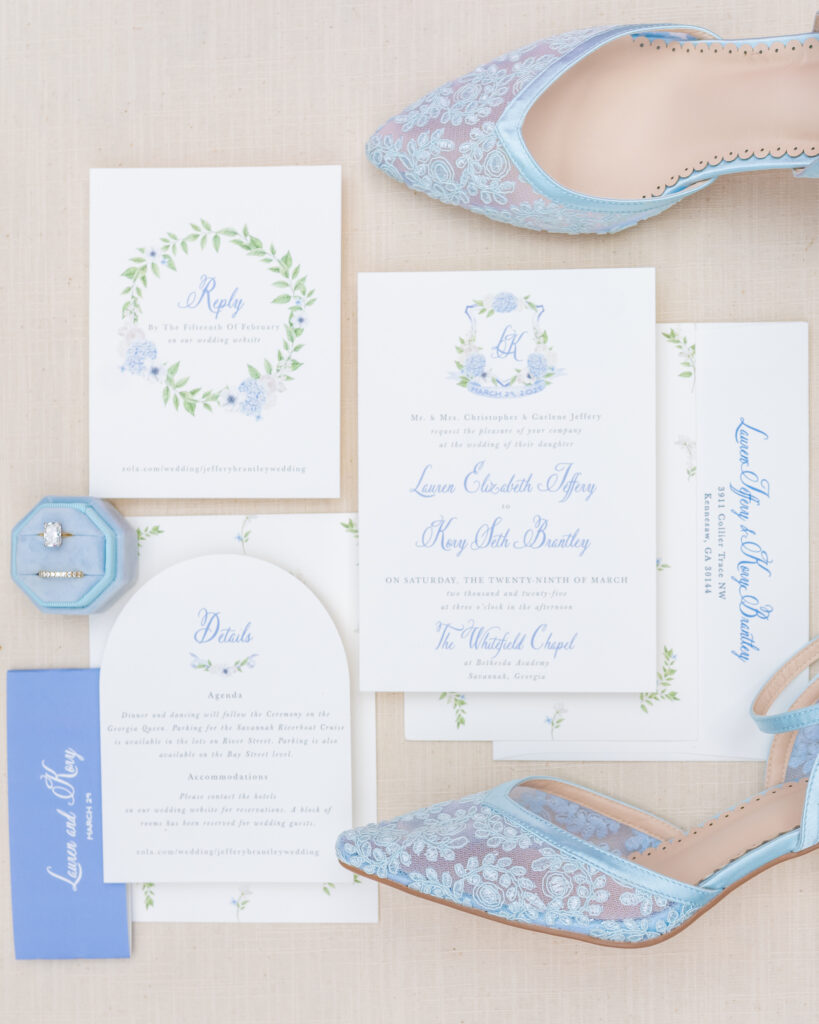 Wedding invitation suite flat lay with a soft blue watercolor crest design, reply card with floral wreath, details card, blue lace bridal shoes, and a blue velvet ring box, photographed by Maryland wedding photographer Jennifer Mary Collective