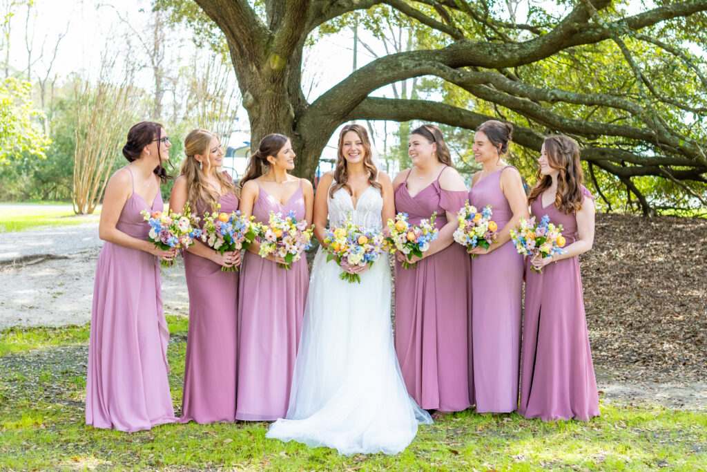 Bride smiling with six bridesmaids in dusty mauve gowns holding colorful wildflower bouquets under a sprawling live oak tree, photographed by Baltimore wedding photographer Jennifer Mary Collective