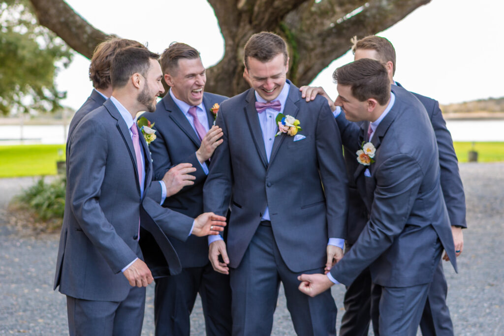 Groomsmen in charcoal suits with pink ties laughing together candidly under a large oak tree at a waterfront wedding venue, photographed by Baltimore wedding photographer Jennifer Mary Collective