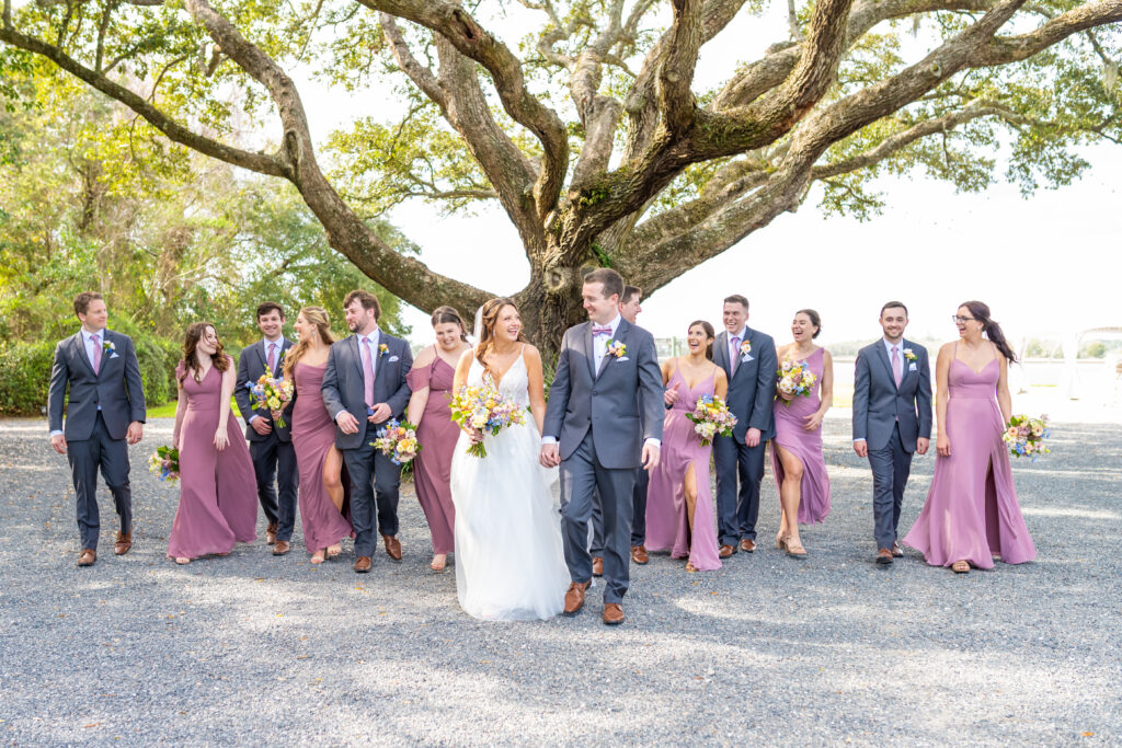 Bride and groom holding hands and smiling while their full wedding party walks behind them under a large live oak tree, photographed by Baltimore wedding photographer Jennifer Mary Collective