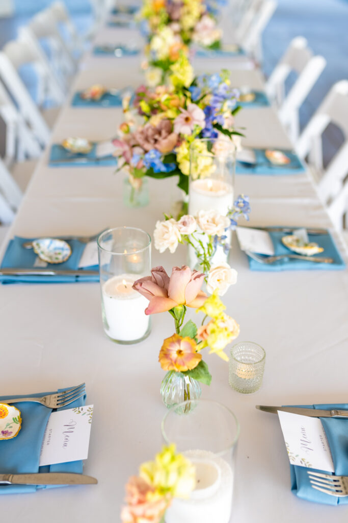 Overhead view of a Maryland wedding reception table with colorful wildflower centerpieces in bud vases, blue linen napkins, floral menu cards, and vintage china, photographed by Maryland wedding photographer Jennifer Mary Collective