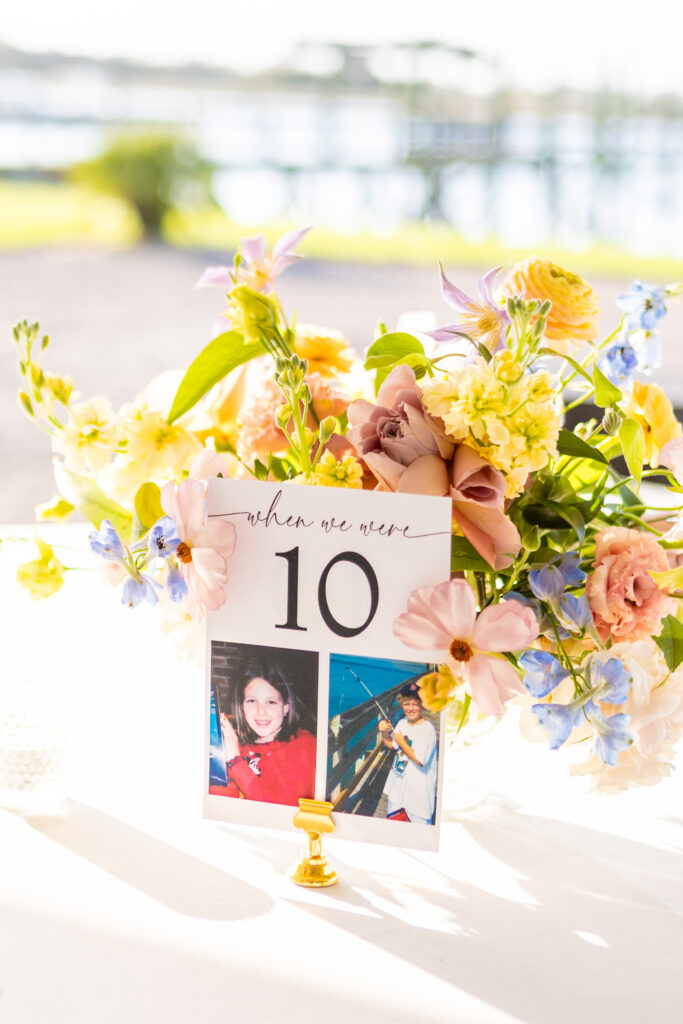 Custom wedding table number featuring childhood photos of the couple beside a lush colorful wildflower centerpiece at a waterfront Maryland wedding reception, photographed by Maryland wedding photographer Jennifer Mary Collective