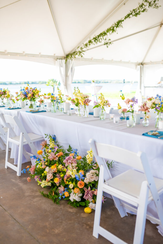 Waterfront Maryland wedding reception inside a white tent with long tables, wildflower centerpieces, and a lush colorful floor floral installation cascading beside the head table, photographed by Maryland wedding photographer Jennifer Mary Collective
