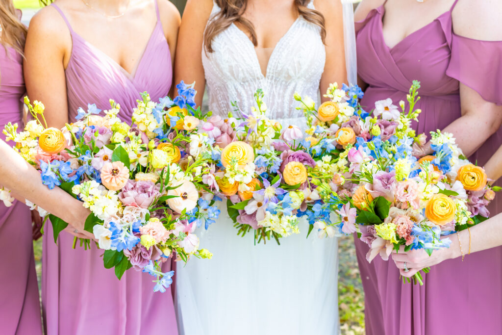 Close up of bride and bridesmaids in mauve dresses holding vibrant wildflower bouquets with orange ranunculus, blue delphinium, blush roses, and lush greenery, photographed by Maryland wedding photographer Jennifer Mary Collective