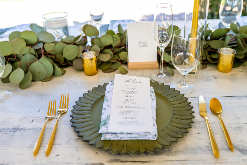 Wedding reception place setting with an olive green sunflower charger plate, gold flatware, printed menu card, and a eucalyptus garland runner at a Maryland waterfront wedding, photographed by Maryland wedding photographer Jennifer Mary Collective