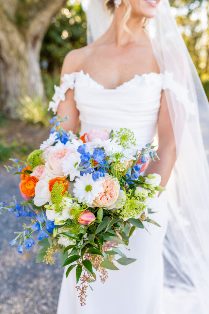 Bride in an off the shoulder gown with a cathedral veil holding a lush colorful bridal bouquet with orange ranunculus, blue delphinium, daisies, and garden roses, photographed by Maryland wedding photographer Jennifer Mary Collective